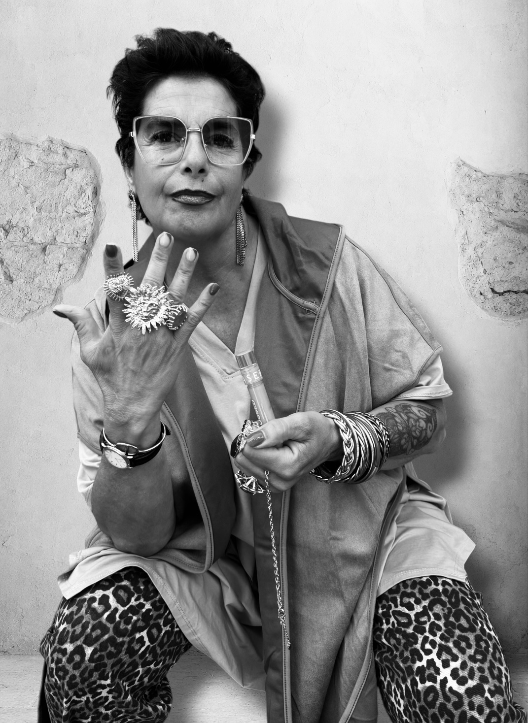 Portrait en noir et blanc d'une femme aux lunettes carrées, veste sans manches en daim, pantalon léopard et bagues argentées, posant avec assurance contre un mur de pierre.